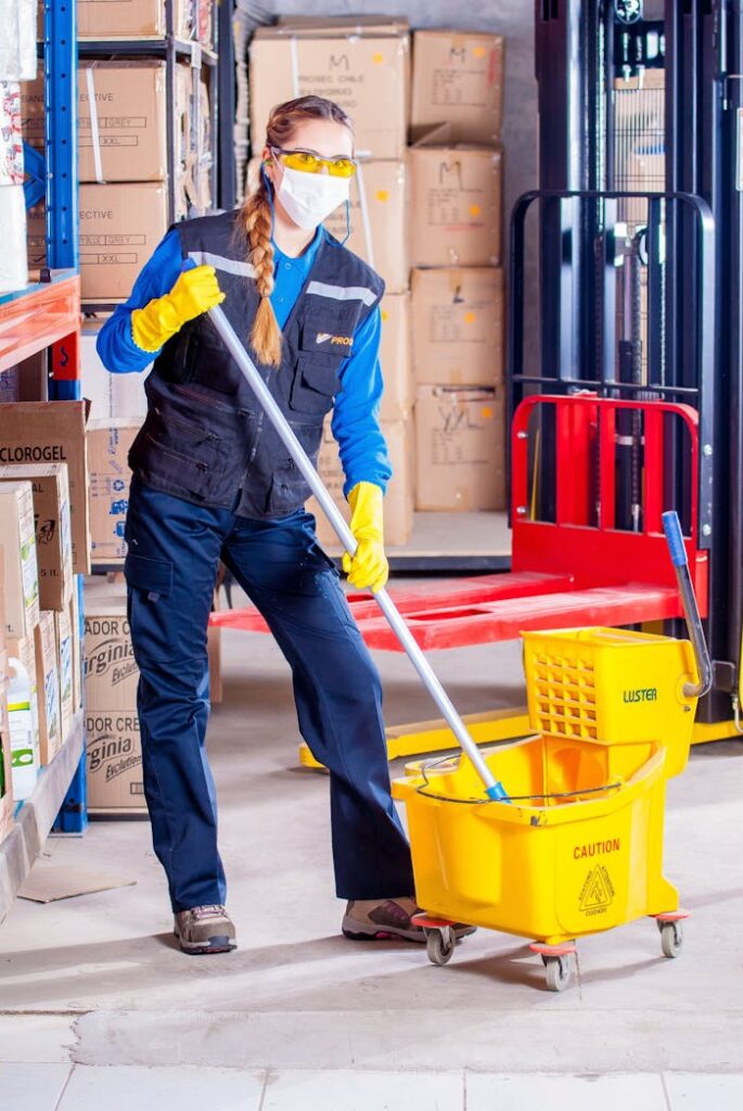 pexels photo 209271 Female janitor in safety gear cleaning a warehouse floor with a mop and bucket.